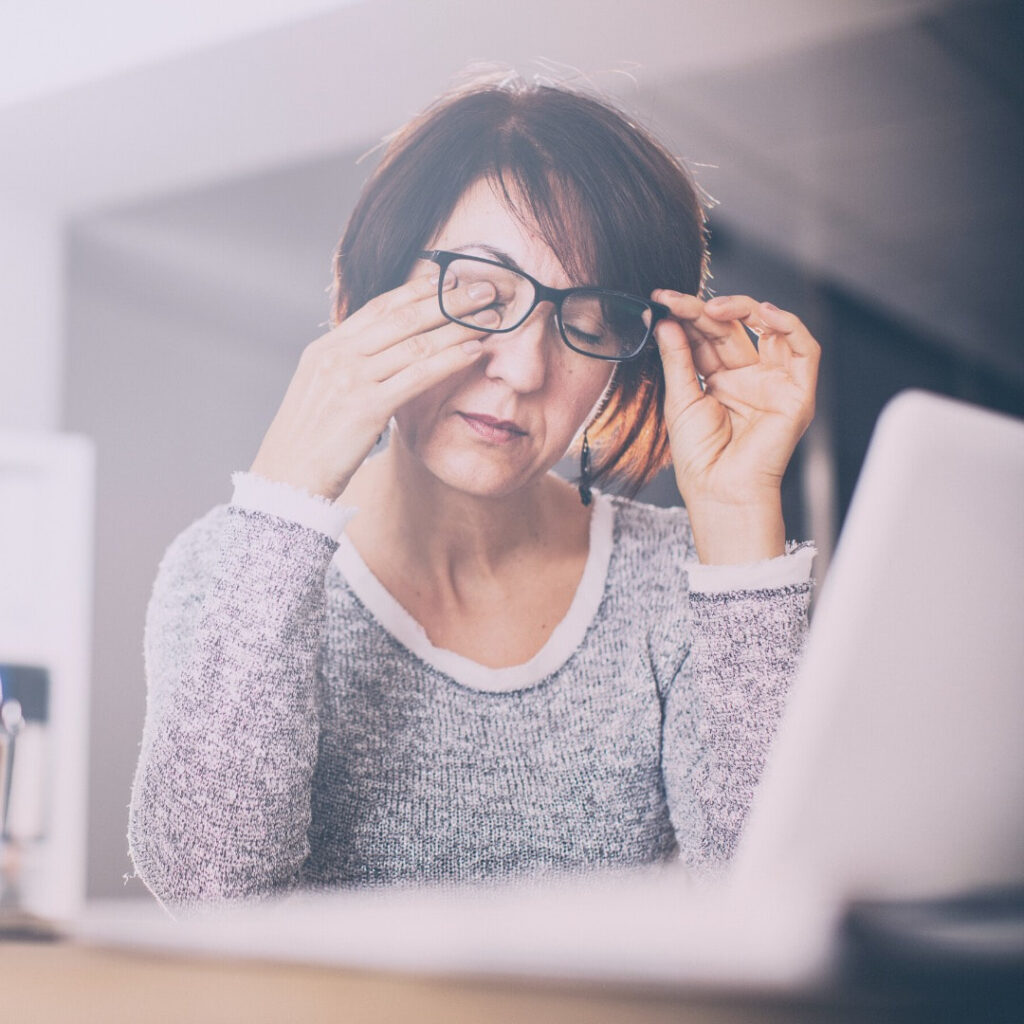 Woman rubbing her eyes and removing her glasses at a desk with laptop—a moment of pause to relax and reset.