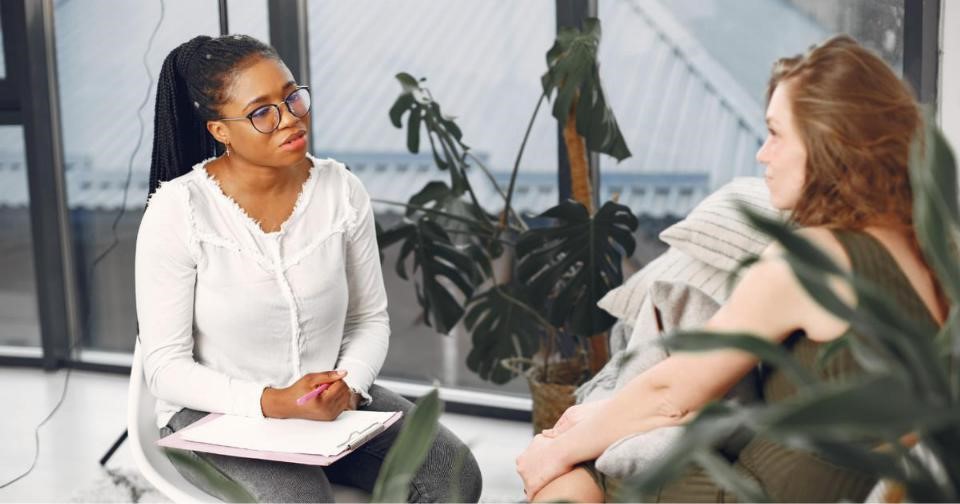 A psychologist in a chair conversing with another woman, while she holds a pen and a notebook.
