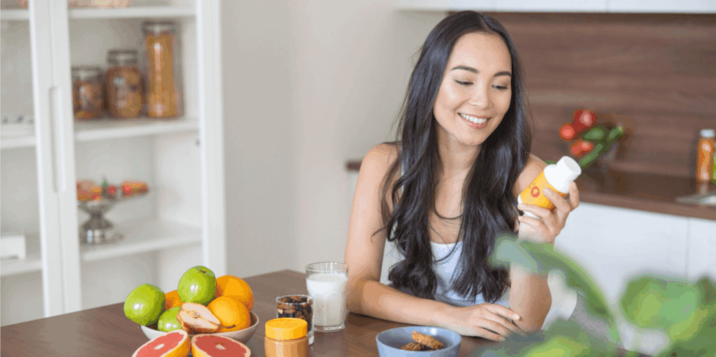 A young woman smiles while holding a container of a supplement, surrounded by fruits, healthy drinks, and vitamins.