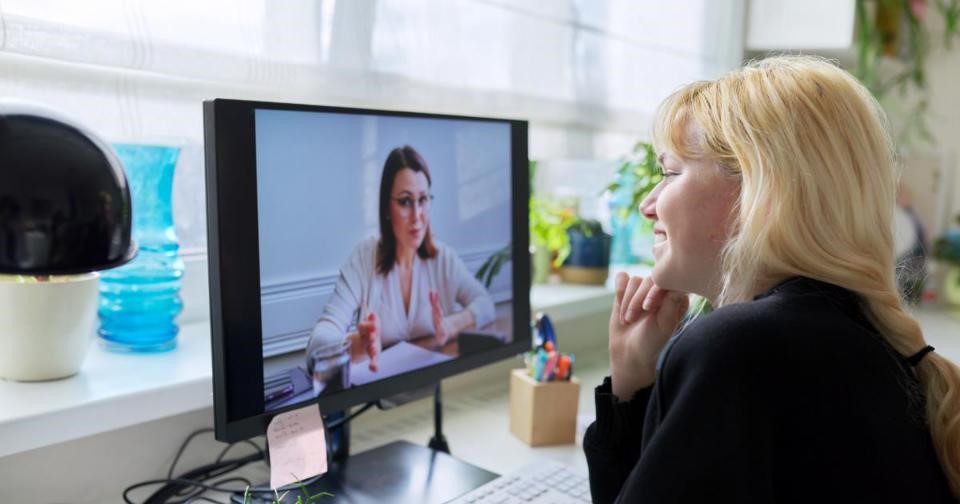 A woman sitting at a desk with a computer screen, engaged in a virtual therapy session with a therapist.