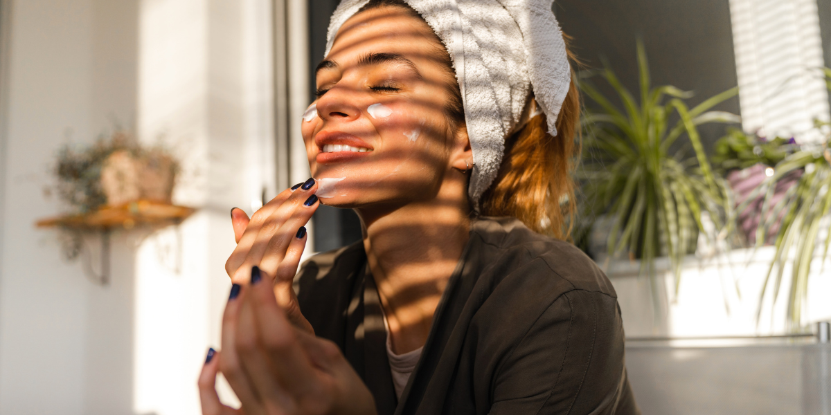 A woman with a towel on her head applying moisturizer as part of her self-care routine.
