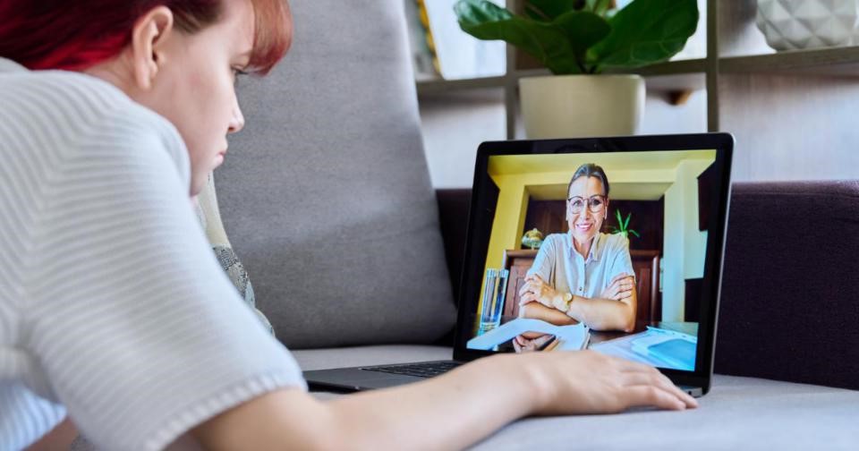 A woman sitting on a couch with a laptop, another  woman on the screen. Image depicts virtual therapy session.
