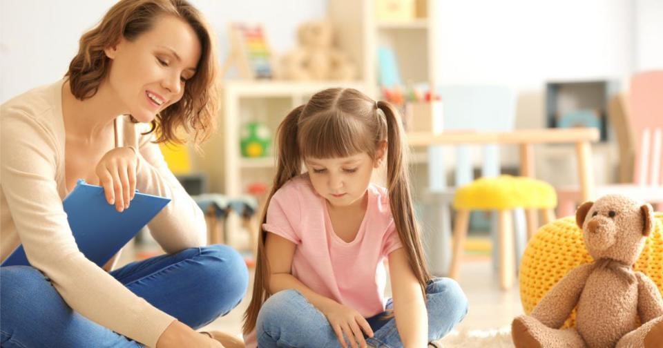 Woman and child sitting on floor with teddy bear during child therapy session.